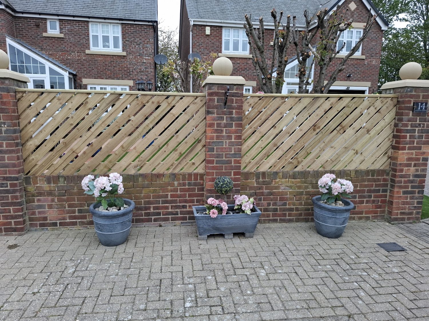 A brick wall topped with diagonal trellis panels above flower pots on block paving