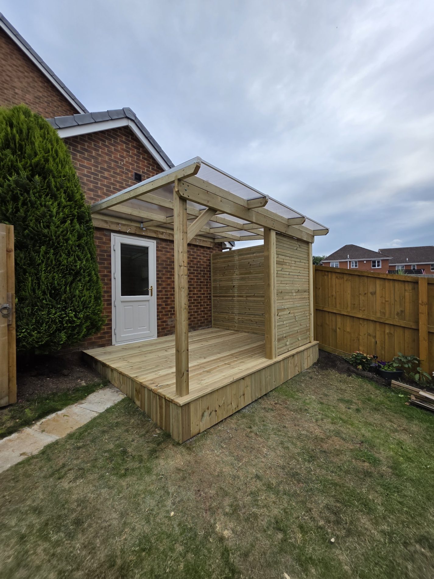 A timber shelter with a clear roof beside a house, on a small deck